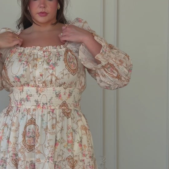 A woman standing in front of a white door wearing a long dress. She has a confident stance and her hair is styled in a neat updo. Her dress is a light color with a pattern of small flowers and