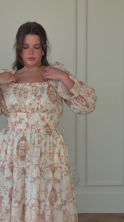 A woman standing in front of a white door wearing a long dress. She has a confident stance and her hair is styled in a neat updo. Her dress is a light color with a pattern of small flowers and
