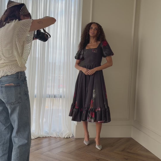 A woman standing in front of a mirror in a room, with a person standing on the left side of the image holding an object in their hand. In the background, there is a curtain and a wall, and at the