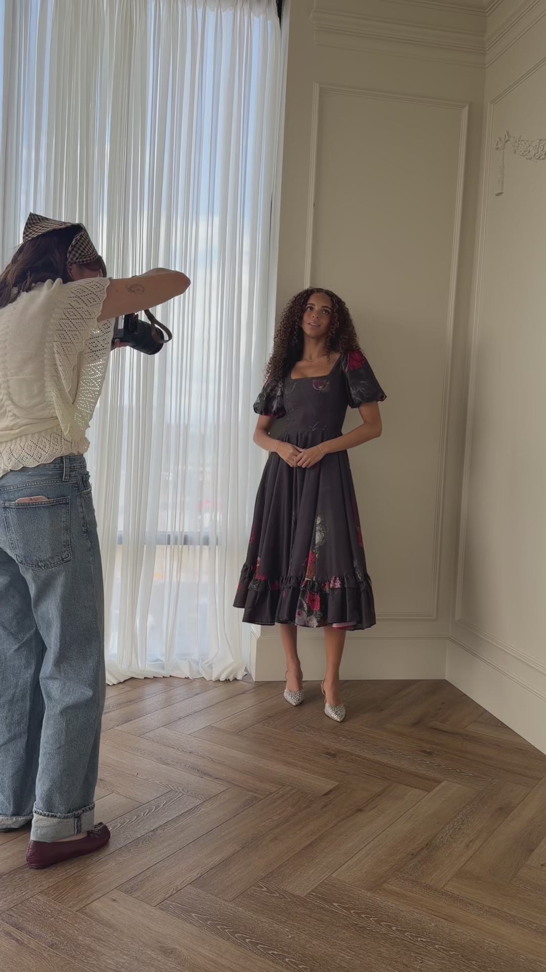 A woman standing in front of a mirror in a room, with a person standing on the left side of the image holding an object in their hand. In the background, there is a curtain and a wall, and at the