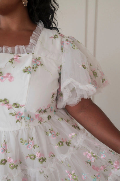 The image shows a woman wearing a white dress with pink flowers on it, standing in front of a wall. The dress has a ruffled hemline and a full skirt that falls to the floor. The flowers are in