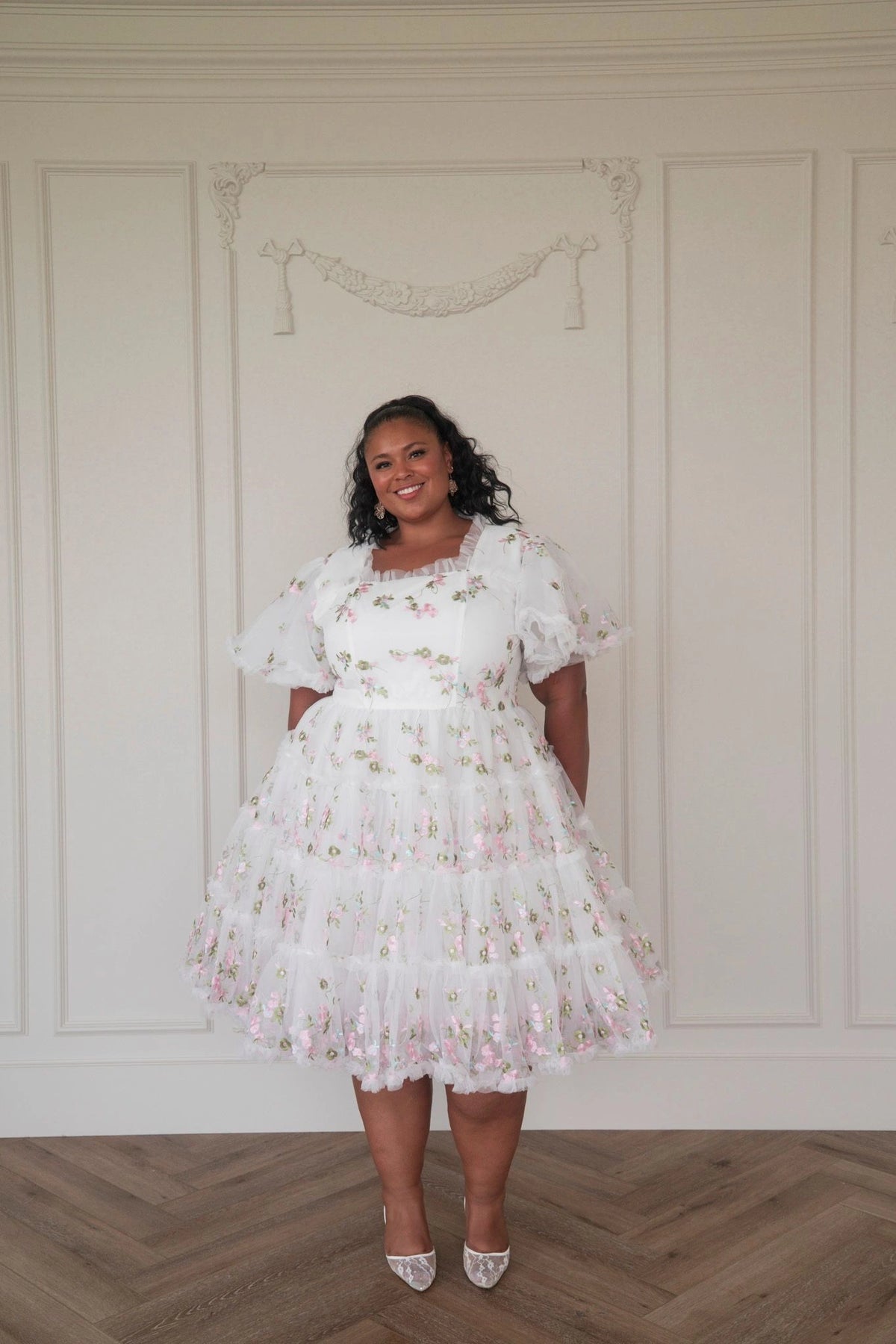 The image shows a woman in a white dress standing in front of a white wall, with a smile on her face. She is wearing a white frock with a pattern of small flowers and leaves, and her hair is styled