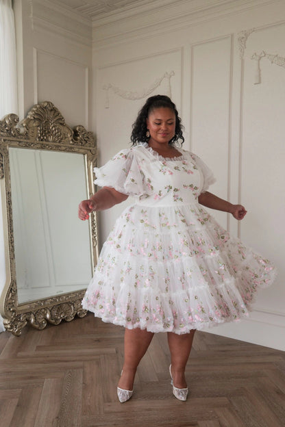 The image shows a woman in a white dress standing in front of a mirror, with a wall in the background. She is wearing a white frock with a pattern of small flowers and leaves, and her hair is styled