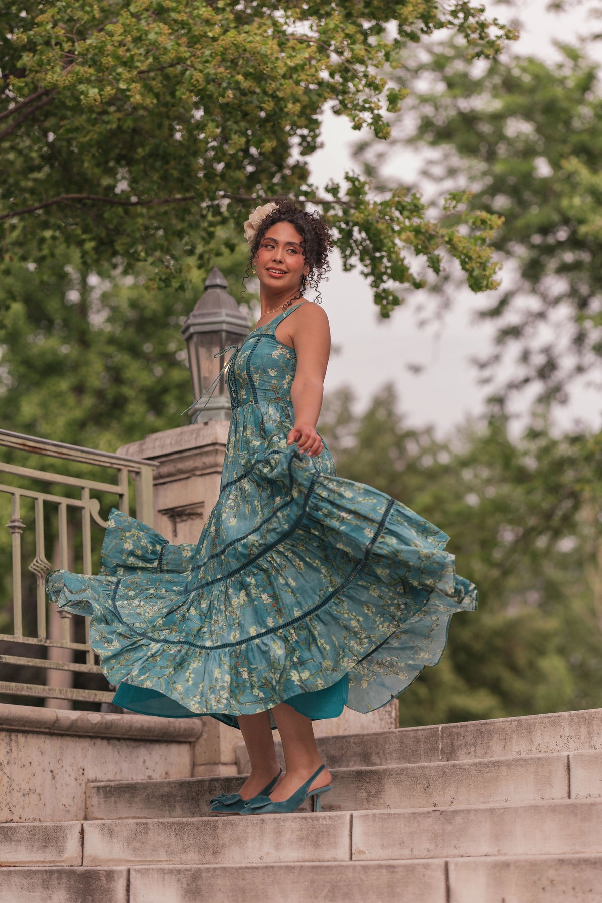 Woman in teal floral dress and flower crown standing on stone steps, smiling at camera with dress flowing outward.